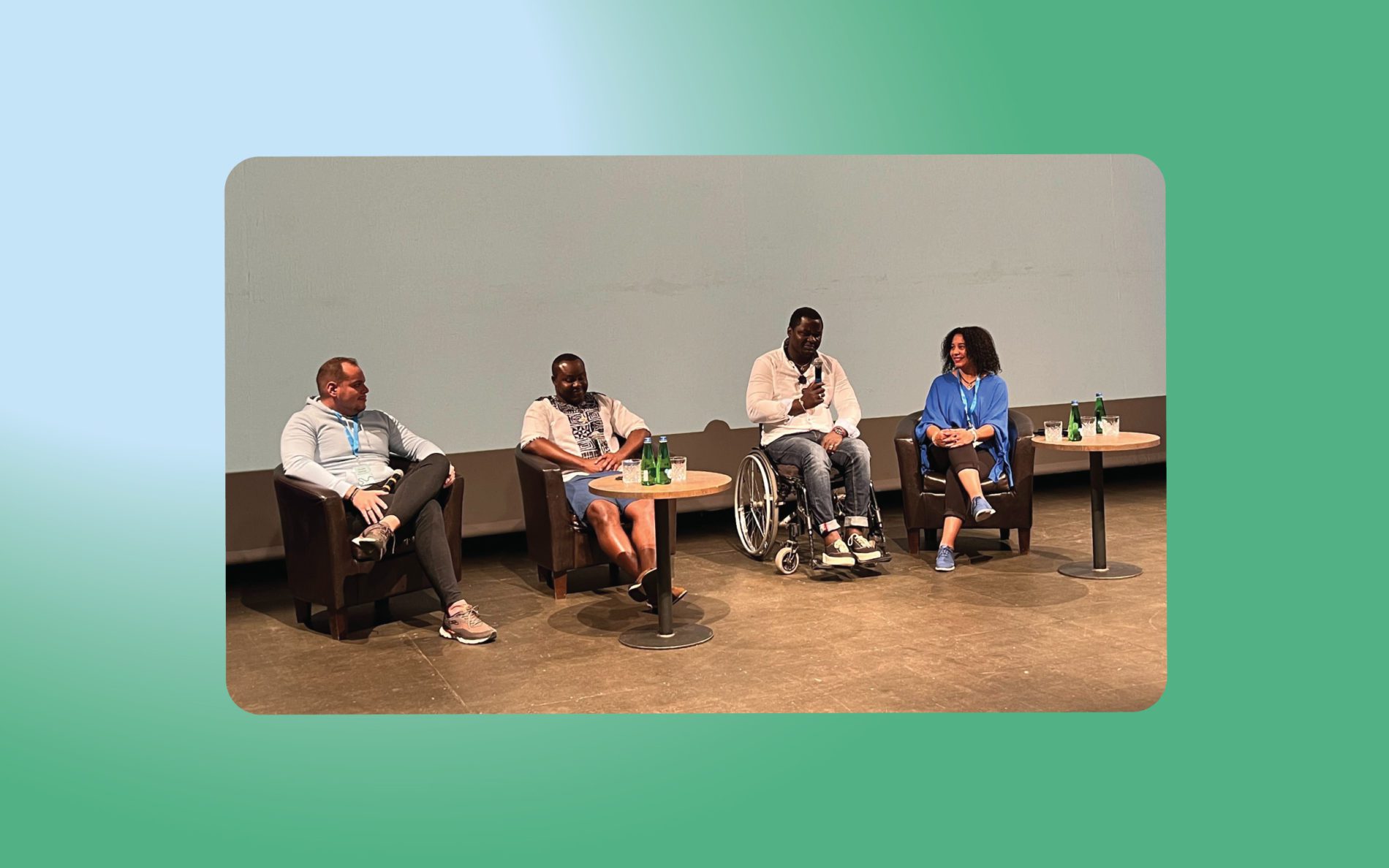 Three people in armchairs and one person in a wheelchair on on the stage, talking.