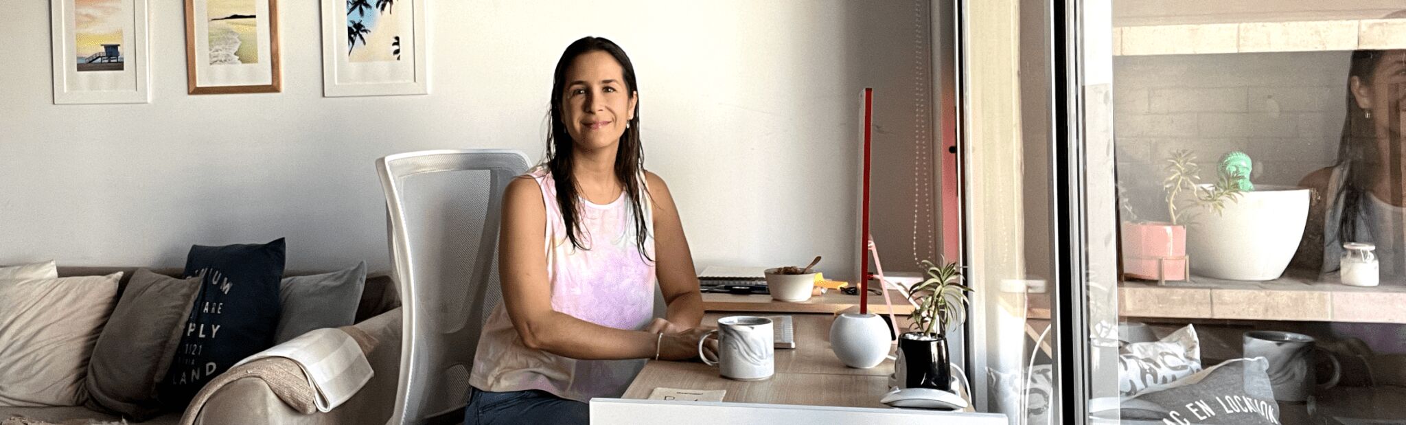 Renata sitting at her desk and smiling at the camera, a coffee cup and laptop are on the desk, there’s a couch with comfy pillows on the right and a...